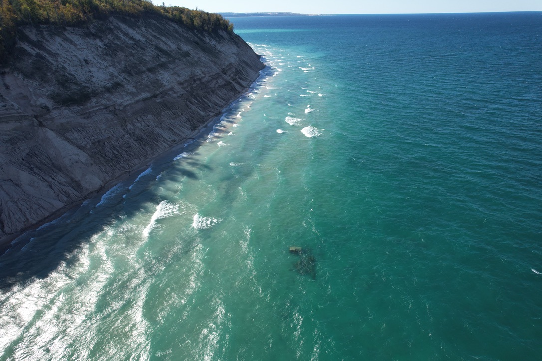 Turquoise water and breaking waves along a steep, tree-lined sand bluff.
